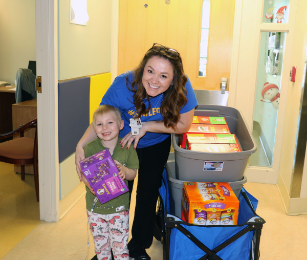 A white blond haired little girls smiles and holds a box of cookies next to a white nurse with brown hair next to a wagon filled with donated snacks like cookies and crackers.