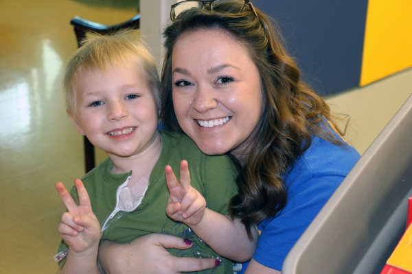 A white blond haired little girls smiles and holds up two peace signs with her fingers as she is in the arms of a white female nurse with brown hair. 